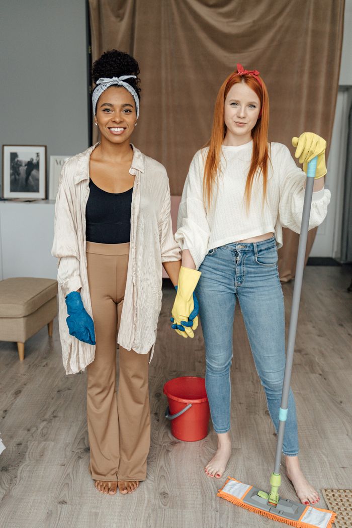 Two women smiling and holding hands while cleaning indoors, symbolizing teamwork.